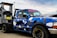 A blue pickup truck with a geometric camouflage pattern is parked next to a diesel fuel pump. The truck has custom stickers and decals on the windshield and body. The fuel pump is labeled 'Marked Diesel'.