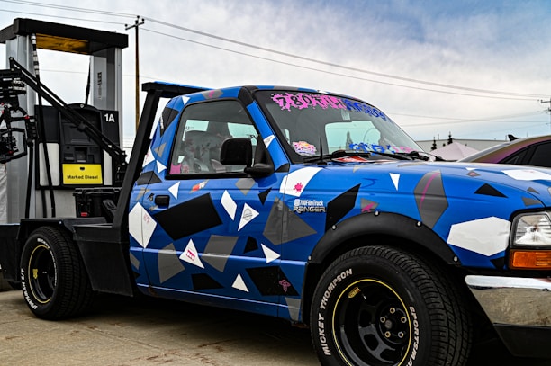 A blue pickup truck with a geometric camouflage pattern is parked next to a diesel fuel pump. The truck has custom stickers and decals on the windshield and body. The fuel pump is labeled 'Marked Diesel'.
