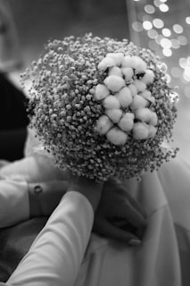 A close-up black and white photograph of a person holding a bouquet of flowers, primarily composed of delicate small buds and cotton-like florets. The image has a bokeh effect in the background with soft circular light patterns.