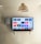 A modern living room features a wall-mounted flat-screen television displaying multiple streaming app icons, such as Apple TV, HBO Max, Netflix, Disney+, and Prime Video. Below the TV is a soundbar sitting on a small white shelf. A wicker pendant light hangs from the ceiling above, and the wall is painted a neutral color.