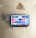 A modern living room features a wall-mounted flat-screen television displaying multiple streaming app icons, such as Apple TV, HBO Max, Netflix, Disney+, and Prime Video. Below the TV is a soundbar sitting on a small white shelf. A wicker pendant light hangs from the ceiling above, and the wall is painted a neutral color.