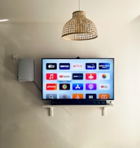 A modern living room features a wall-mounted flat-screen television displaying multiple streaming app icons, such as Apple TV, HBO Max, Netflix, Disney+, and Prime Video. Below the TV is a soundbar sitting on a small white shelf. A wicker pendant light hangs from the ceiling above, and the wall is painted a neutral color.