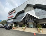 A modern, geometric building with reflective glass and metal panels. Several business signs are prominently displayed. The entrance features large, angular awnings and a staircase leading up to glass doors. There are several cars parked in front, a few people standing near the vehicles, and decorative plants in large pots lining the stairs.