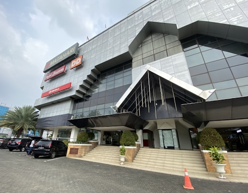 A modern, geometric building with reflective glass and metal panels. Several business signs are prominently displayed. The entrance features large, angular awnings and a staircase leading up to glass doors. There are several cars parked in front, a few people standing near the vehicles, and decorative plants in large pots lining the stairs.