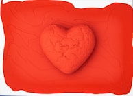 A textured heart made from clay or a similar material is positioned in the center of a vibrant red background. The heart appears slightly cracked and uneven, adding an artistic, handmade quality to the image. The background is smooth but has some subtle patterns and contours, which enhance the heart's prominence.