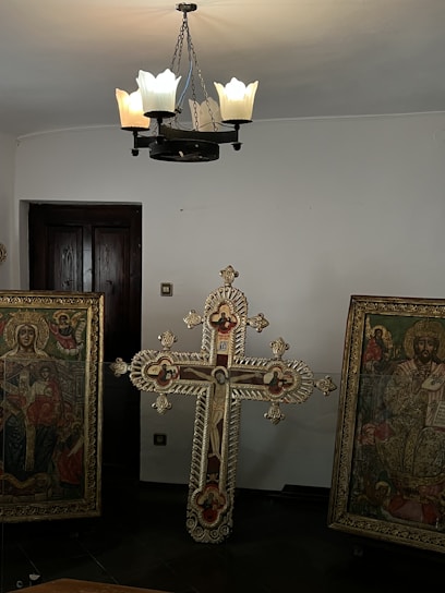 The image features a room with a decorative chandelier hanging from the ceiling, emitting soft light. Below, there are religious artifacts, including an ornately designed cross and two framed religious paintings depicting iconic Christian figures in detailed and vivid colors. The walls and door in the background are plain, providing contrast to the intricate designs of the artifacts.