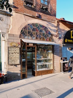 A small storefront with a vintage design featuring a variety of jewelry displayed in the window. The shop has a decorative arch window above the entrance, with iron bars on the windows upstairs. To the right, there is another business with a sign indicating it is a travel agency. A person is walking by on the sidewalk, and a street sign with different types of keys is visible on the left side.