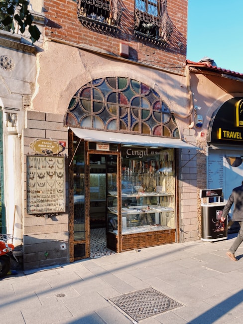 A small storefront with a vintage design featuring a variety of jewelry displayed in the window. The shop has a decorative arch window above the entrance, with iron bars on the windows upstairs. To the right, there is another business with a sign indicating it is a travel agency. A person is walking by on the sidewalk, and a street sign with different types of keys is visible on the left side.