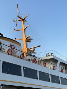 A large ferry with a prominent yellow mast and several lifebuoys attached to the railing is docked. The upper part of the vessel features multiple windows and safety equipment. The clear blue sky enhances the bright appearance of the ship.