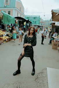 A woman in a dark dress and black shoes stands confidently in the center of a busy outdoor market. Around her, people shop at various stalls filled with clothing and fresh produce. The buildings in the background have a pastel color scheme with mint green roofs.