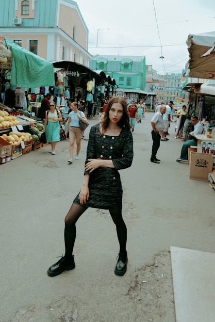 A woman in a dark dress and black shoes stands confidently in the center of a busy outdoor market. Around her, people shop at various stalls filled with clothing and fresh produce. The buildings in the background have a pastel color scheme with mint green roofs.