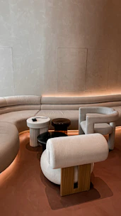 A modern interior with a curved sofa set against a textured beige wall, accompanied by a collection of uniquely shaped chairs and small round tables. The lighting is soft and ambient, creating an inviting atmosphere.