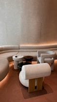 A modern interior with a curved sofa set against a textured beige wall, accompanied by a collection of uniquely shaped chairs and small round tables. The lighting is soft and ambient, creating an inviting atmosphere.