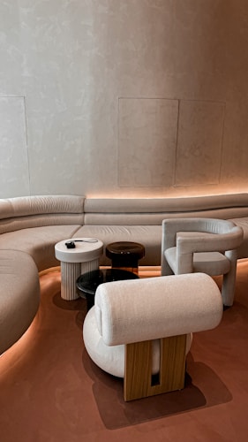 A modern interior with a curved sofa set against a textured beige wall, accompanied by a collection of uniquely shaped chairs and small round tables. The lighting is soft and ambient, creating an inviting atmosphere.