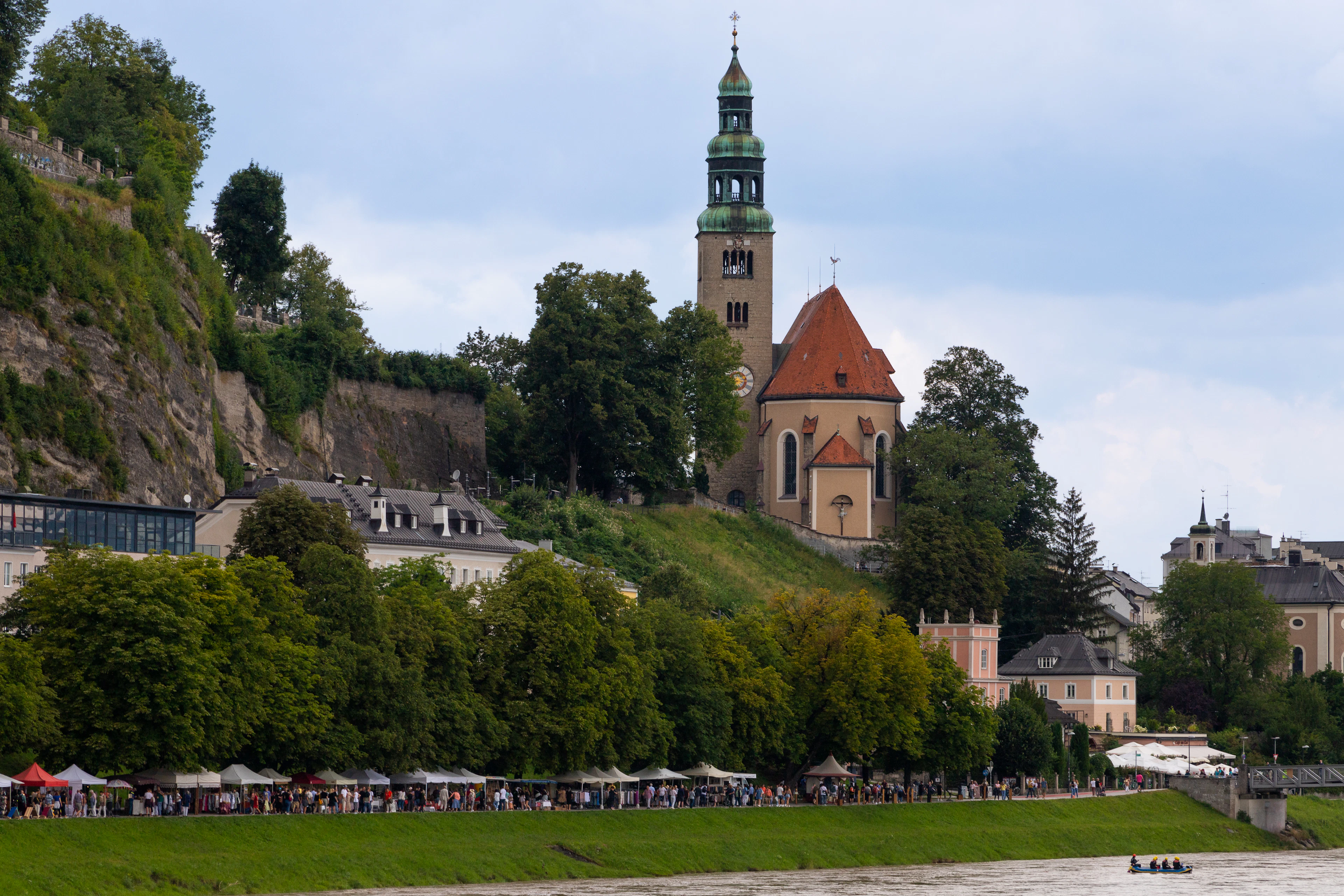 The green onion-domed bell tower and red-tiled nave of the Mulln parish church standing on the wooded Monchsberg bluff above the Salzach in Salzburg, with white market canopies and strollers along the grassy riverbank below.