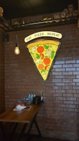 A large slice of pizza is depicted in a neon sign on a brick wall, with toppings like pepperoni, mushrooms, and leaves. Below the sign, there is a wooden table with several items, including water bottles, stacked plates, and condiments. The setting is illuminated by a hanging light bulb, creating a cozy atmosphere.