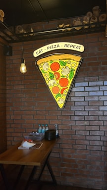 A large slice of pizza is depicted in a neon sign on a brick wall, with toppings like pepperoni, mushrooms, and leaves. Below the sign, there is a wooden table with several items, including water bottles, stacked plates, and condiments. The setting is illuminated by a hanging light bulb, creating a cozy atmosphere.