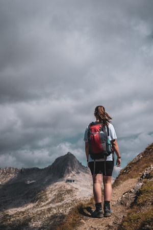 A person stands on a narrow hiking trail, facing away, with a prominent mountain peak in the background. The individual is wearing outdoor clothing and carrying a red backpack, suggesting a hiking or trekking activity. The sky is overcast with thick clouds, creating a dramatic atmosphere.