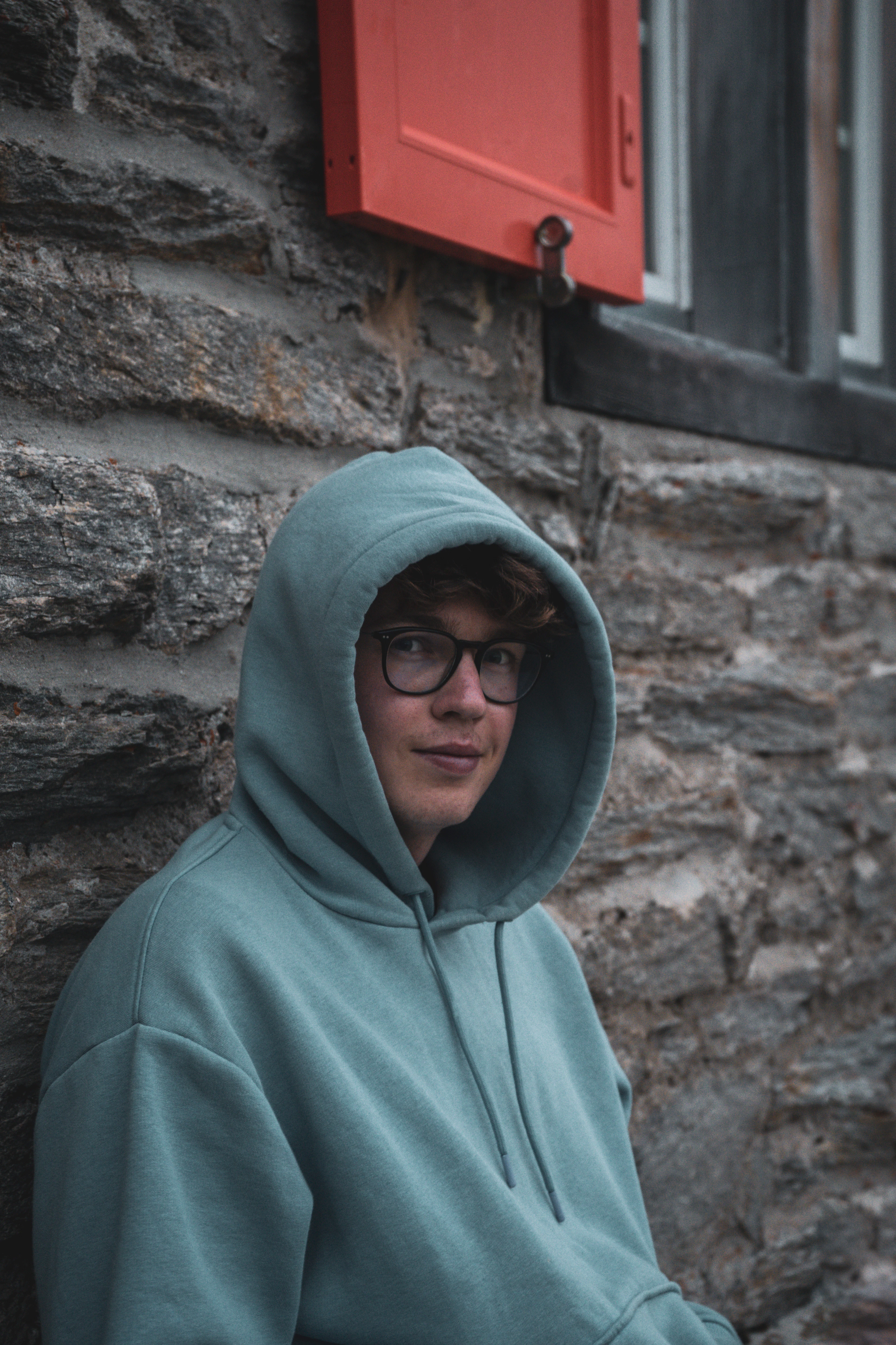 A young person in a sea-green hoodie leaning against a stone wall.