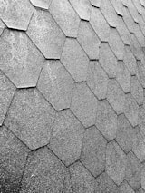 A close-up view of a roof with hexagonal asphalt shingles arranged in a repeating pattern. The texture is rough and appears consistent across the surface.