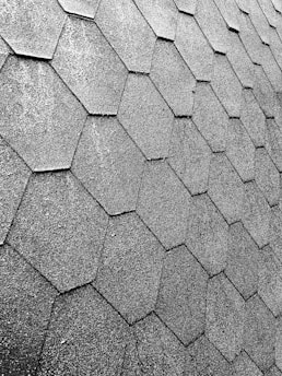 A close-up view of a roof with hexagonal asphalt shingles arranged in a repeating pattern. The texture is rough and appears consistent across the surface.