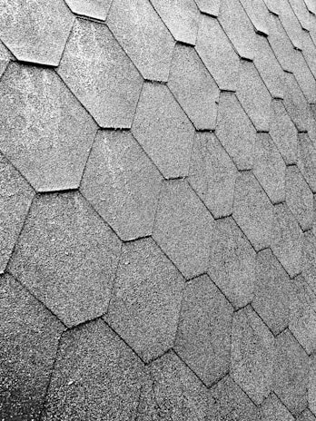 A close-up view of a roof with hexagonal asphalt shingles arranged in a repeating pattern. The texture is rough and appears consistent across the surface.