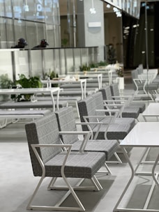 A modern and sleek seating area with rows of geometric patterned chairs and white metal frames, lined up alongside rectangular tables. The ambiance appears minimalistic, with clean lines and a bright, airy feel. The background includes a glass partition with potted plants in a well-lit space.