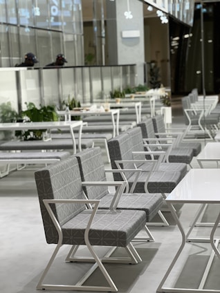 A modern and sleek seating area with rows of geometric patterned chairs and white metal frames, lined up alongside rectangular tables. The ambiance appears minimalistic, with clean lines and a bright, airy feel. The background includes a glass partition with potted plants in a well-lit space.