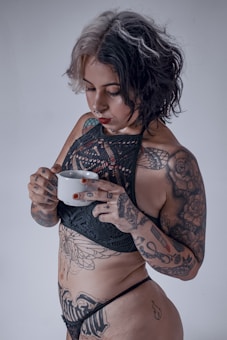 A person with multiple tattoos on their body holds a white cup. They have short hair with a mix of black and gray colors and are wearing a black lace bralette and underwear. The tattoos include floral and script designs.