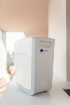 A modern reverse osmosis water filtration device, primarily white and gray in color, is placed on a clean, light-colored surface. The brand name 'Bluewater' and a blue circular button with an 'O' are visible on the front. The device has smooth and sleek contours, indicating a minimalist and contemporary design.