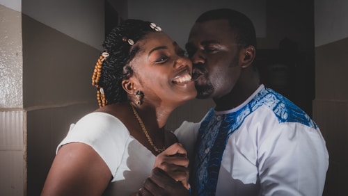 A joyful couple shares a tender moment, with a man affectionately kissing the woman's cheek. They both appear to be dressed in elegant attire, with her wearing a white dress and his shirt featuring blue and white patterns. The woman's hair is styled with beads, adding to the festive or celebratory feel.