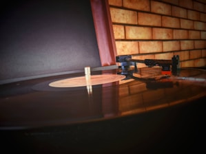 A close-up view of a vintage record player with a vinyl record on the turntable. The focus is on the tonearm and stylus poised over the record. The background is a textured brick wall, creating a nostalgic atmosphere.