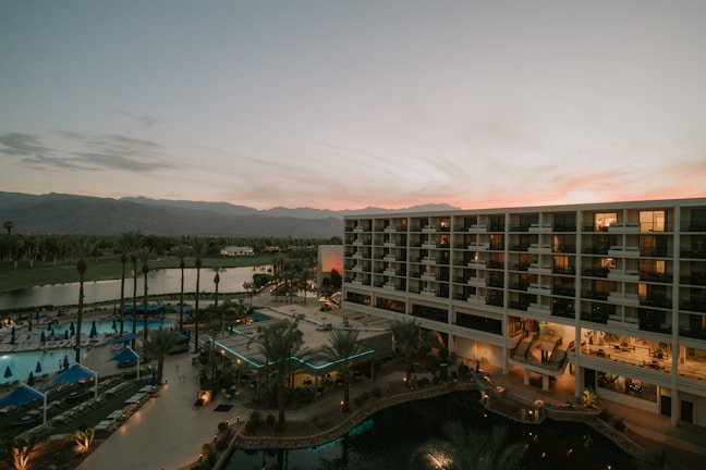 An overview of a resort or hotel complex with a large modern building featuring numerous balconies. In the foreground, there are palm trees and a swimming pool area with lounge chairs and blue umbrellas. The background shows a scenic view of mountains under a sky at sunset, displaying a gradient of pink and orange hues.
