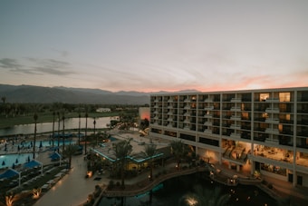 An overview of a resort or hotel complex with a large modern building featuring numerous balconies. In the foreground, there are palm trees and a swimming pool area with lounge chairs and blue umbrellas. The background shows a scenic view of mountains under a sky at sunset, displaying a gradient of pink and orange hues.