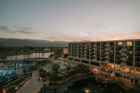 An overview of a resort or hotel complex with a large modern building featuring numerous balconies. In the foreground, there are palm trees and a swimming pool area with lounge chairs and blue umbrellas. The background shows a scenic view of mountains under a sky at sunset, displaying a gradient of pink and orange hues.