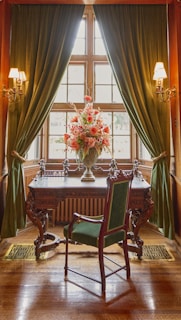 An elegant room features a vintage wooden desk with intricate carvings, positioned in front of a bright window flanked by lush green curtains. On the desk, there is a large ornate vase filled with a vibrant arrangement of pink and red flowers. A tall, antique chair with green upholstery complements the regal setting. Warm light from wall sconces enhances the sophisticated atmosphere.
