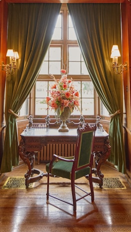 An elegant room features a vintage wooden desk with intricate carvings, positioned in front of a bright window flanked by lush green curtains. On the desk, there is a large ornate vase filled with a vibrant arrangement of pink and red flowers. A tall, antique chair with green upholstery complements the regal setting. Warm light from wall sconces enhances the sophisticated atmosphere.