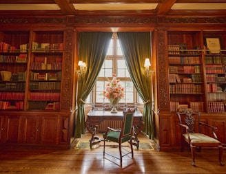 A beautifully furnished library with ornate wooden bookshelves and a wooden desk near a large window. Books of various colors are neatly arranged on the shelves. Two elegant chairs are positioned near the desk, and vibrant flower arrangements are placed in front of green velvet curtains.
