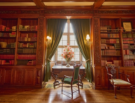 A beautifully furnished library with ornate wooden bookshelves and a wooden desk near a large window. Books of various colors are neatly arranged on the shelves. Two elegant chairs are positioned near the desk, and vibrant flower arrangements are placed in front of green velvet curtains.