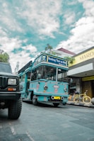 A bright blue tour bus labeled 'Bandung Tour on Bus' is parked on a cobblestone street. There is a yellow bicycle next to a shop with a sign reading 'Mardika.' The sky is partly cloudy, and another vehicle is partially visible in the foreground.