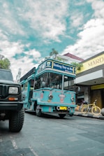 A bright blue tour bus labeled 'Bandung Tour on Bus' is parked on a cobblestone street. There is a yellow bicycle next to a shop with a sign reading 'Mardika.' The sky is partly cloudy, and another vehicle is partially visible in the foreground.