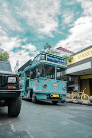 A bright blue tour bus labeled 'Bandung Tour on Bus' is parked on a cobblestone street. There is a yellow bicycle next to a shop with a sign reading 'Mardika.' The sky is partly cloudy, and another vehicle is partially visible in the foreground.