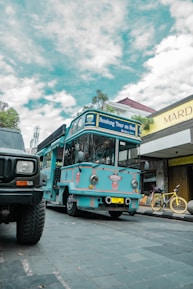 A bright blue tour bus labeled 'Bandung Tour on Bus' is parked on a cobblestone street. There is a yellow bicycle next to a shop with a sign reading 'Mardika.' The sky is partly cloudy, and another vehicle is partially visible in the foreground.