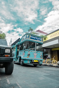 A bright blue tour bus labeled 'Bandung Tour on Bus' is parked on a cobblestone street. There is a yellow bicycle next to a shop with a sign reading 'Mardika.' The sky is partly cloudy, and another vehicle is partially visible in the foreground.