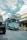 A bright blue tour bus labeled 'Bandung Tour on Bus' is parked on a cobblestone street. There is a yellow bicycle next to a shop with a sign reading 'Mardika.' The sky is partly cloudy, and another vehicle is partially visible in the foreground.