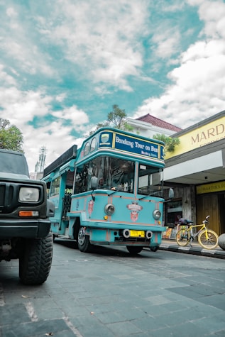 A bright blue tour bus labeled 'Bandung Tour on Bus' is parked on a cobblestone street. There is a yellow bicycle next to a shop with a sign reading 'Mardika.' The sky is partly cloudy, and another vehicle is partially visible in the foreground.
