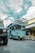 A bright blue tour bus labeled 'Bandung Tour on Bus' is parked on a cobblestone street. There is a yellow bicycle next to a shop with a sign reading 'Mardika.' The sky is partly cloudy, and another vehicle is partially visible in the foreground.