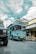 A bright blue tour bus labeled 'Bandung Tour on Bus' is parked on a cobblestone street. There is a yellow bicycle next to a shop with a sign reading 'Mardika.' The sky is partly cloudy, and another vehicle is partially visible in the foreground.