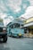 A bright blue tour bus labeled 'Bandung Tour on Bus' is parked on a cobblestone street. There is a yellow bicycle next to a shop with a sign reading 'Mardika.' The sky is partly cloudy, and another vehicle is partially visible in the foreground.