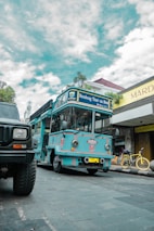 A bright blue tour bus labeled 'Bandung Tour on Bus' is parked on a cobblestone street. There is a yellow bicycle next to a shop with a sign reading 'Mardika.' The sky is partly cloudy, and another vehicle is partially visible in the foreground.